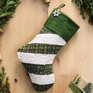 May include: A quilted Christmas stocking featuring alternating horizontal stripes of solid white fabric and dark green fabric with a plaid pattern and small white snowflake motifs. The cuff is solid dark green with a white wooden snowflake ornament.