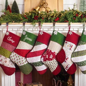 Puede incluir: Seis calcetines navideños acolchados colgados en fila, con rayas diagonales alternas de tela roja, verde y blanca con varios patrones. Dos calcetines están personalizados con los nombres bordados Claire y James.