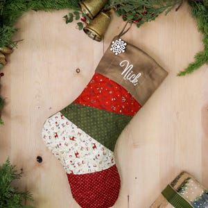 以下が含まれることがあります： 茶色、緑、赤、白のパッチワークのクリスマスストッキングで、カフに「ニック」という名前が刺繍されています。ストッキングは雪の結晶で飾られており、木製の表面の上に置かれています。