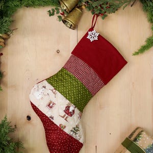 以下が含まれることがあります： 雪だるま、家、サンタクロースが描かれたパッチワークのデザインの赤と緑のクリスマスストッキング。ストッキングの上部には白い雪の結晶のオーナメントが付いています。