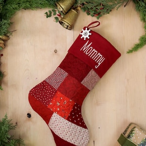 以下が含まれることがあります： 雪の結晶のオーナメントと「Mommy」という刺繍が施された赤いキルティングのクリスマスストッキング