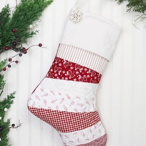 Puede incluir: Una media de Navidad acolchada roja y blanca con un adorno de copo de nieve. La media está hecha de diferentes telas con varios patrones navideños, incluyendo muñecos de nieve, árboles y cuadros.