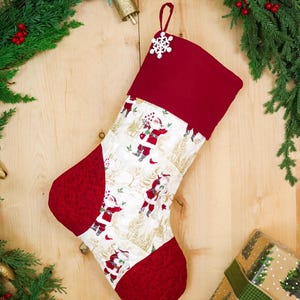 Op de afbeelding: Een kerstkous met een wit stoffen middenstuk met een herhalend patroon van kerstmanfiguren en gouden bomen. De boord, hiel en teen zijn gemaakt van effen donkerrode stof. Een kleine witte sneeuwvlokversiering is aan de bovenste boord bevestigd.