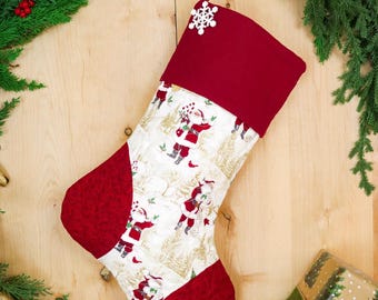 Pode incluir: Uma meia de Natal com um centro de tecido branco apresentando um padrão repetitivo de figuras do Pai Natal e árvores douradas. O punho, o calcanhar e a biqueira são feitos de tecido vermelho escuro sólido. Um pequeno enfeite de floco de neve branco está preso ao punho superior.