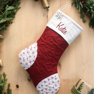 以下が含まれることがあります： 赤色の本体と白い袖口が付いた、キルティング加工のクリスマスストッキング。「Katie」という名前が赤い文字で刺繍されています。ストッキングには雪の結晶のデザインと雪の結晶のチャームが付いています。