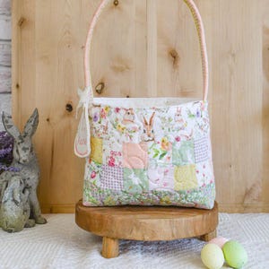 Może przedstawiać: Pikowany koszyk wielkanocny z różowym uchwytem i patchworkowym wzorem z króliczkami i motywami kwiatowymi. Koszyk stoi na małym drewnianym stojaku, w pobliżu znajdują się dekoracyjne pisanki. Ogólna kolorystyka jest pastelowa.