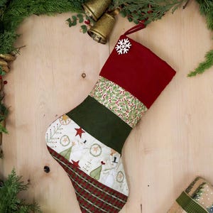 以下が含まれることがあります： 雪の結晶のオーナメントが付いた赤と緑のクリスマスストッキング。ストッキングは、雪だるま、クリスマスツリー、赤い星が描かれたパッチワークのデザインで飾られています。