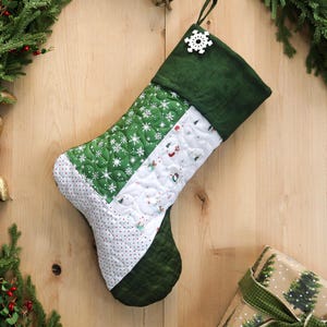 以下が含まれることがあります： 緑色の袖口と雪の結晶のボタンが付いた、キルト加工のクリスマスストッキング。ストッキングは、緑色の雪の結晶と赤いドット模様のパッチワークデザインが特徴です。緑のリボンで包まれたギフトが隅にあります。