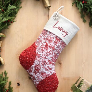 以下が含まれることがあります： 赤い文字で「Lainey」と刺繍された白いカフが付いたクリスマスストッキング。ストッキングの本体は赤と白のトワルドジュイ柄で、赤いキルトのアクセントが付いています。ループには雪の結晶のチャームがぶら下がっています。