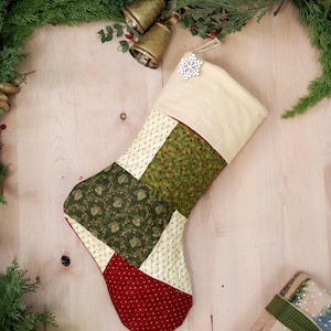 以下が含まれることがあります： 緑、赤、クリーム色のパッチワークのクリスマスストッキングに、雪の結晶の装飾が施されています。ストッキングは、緑色のガーランドと金のベルが飾られた木製の表面に吊り下げられています。