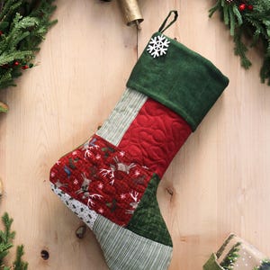 以下が含まれることがあります： 緑色の袖口と白い雪の結晶のアクセントが付いた、キルティング加工のクリスマスストッキング。本体は、赤い生地、トナカイ、白い雪の結晶を使ったパッチワークデザインです。つま先には緑と白のストライプ生地を使用しています。