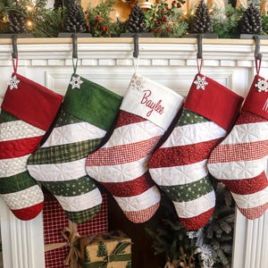以下が含まれることがあります： 暖炉に飾られた5つのクリスマスストッキング。ストッキングは、ストライプ、チェック、水玉模様など、赤、緑、白の模様が特徴です。「Baylee」と「Nicole」という名前が刺繍されたストッキングが2つあります。