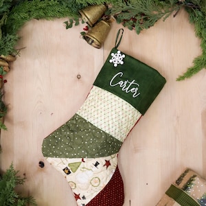 以下が含まれることがあります： 雪の結晶のオーナメントとカフに「Carter」という名前が刺繍された、緑、赤、クリーム色のキルティングされたクリスマスストッキングです。