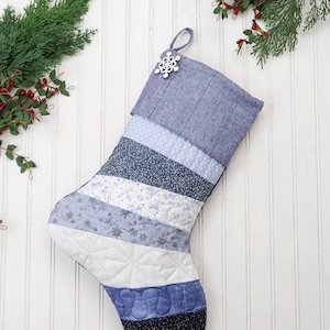 以下が含まれることがあります： 雪の結晶のオーナメントが付いた青と白のキルティングのクリスマスストッキング。ストッキングは、雪の結晶模様のさまざまな青と白の生地で作られています。