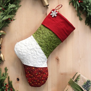 以下が含まれることがあります： 赤、緑、白のセクションがあるキルトのクリスマスストッキング。上部は赤、中央は緑、下部は赤い模様の白です。赤いループから雪の結晶のチャームがぶら下がっています。お祝いの休日の装飾。