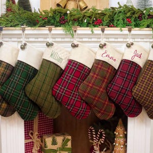 Puede incluir: Siete calcetines navideños colgados en fila, cada uno con un puño color crema y un patrón de tela a cuadros único en tonos verde, rojo, marrón y beige. Algunos calcetines están personalizados con nombres bordados como William, Emma, Jack y Kaitlyn.