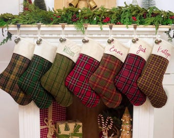 May include: Seven Christmas stockings hanging in a row, each featuring a cream-coloured cuff and a unique plaid fabric pattern in shades of green, red, brown, and tan. Some stockings are personalised with embroidered names including William, Emma, Jack, and Kaitlyn.