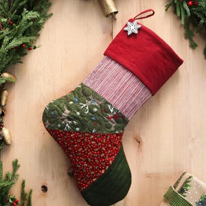 以下が含まれることがあります： 赤いカフと雪の結晶の飾りが付いた、キルティング加工のクリスマスストッキング。赤と白のストライプ生地、鹿と鳥の模様の緑色の生地、白いドットの赤い生地が使用されています。かかとは緑色です。
