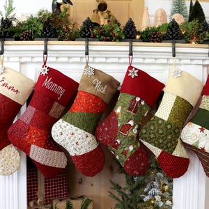 以下が含まれることがあります： 白い暖炉の棚に、パーソナライズされたクリスマスストッキングが5つ吊るされています。ストッキングは、赤、緑、クリーム色の生地でできており、パッチワークのデザインになっています。ストッキングには、「Emma」、「Mommy」、「Nick」という名前と、画像では見えない他の2つの名前が刺繍されています。