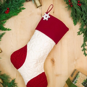 May include: A Christmas stocking featuring a cream-coloured quilted body with a swirling pattern, accented by a deep red cuff, heel, and toe. A white snowflake ornament with a central red gem is attached to the top loop for hanging.
