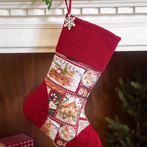 Op de afbeelding: Een kerstkous met een rode boord en teen met een doorgestikte textuur. Het middenpaneel toont een patchworkprint van peperkoekhuisjes, vrachtwagens en koekjes met de tekst Gingerbread Baking Co. Een witte houten sneeuwvlokversiering hangt aan de bovenkant.