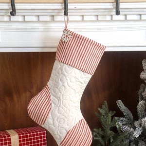 Puede incluir: Un calcetín navideño acolchado con cuerpo blanco y detalles a rayas rojas y blancas. La parte superior y el talón del calcetín presentan rayas, mientras que el cuerpo principal tiene un patrón acolchado. Un copo de nieve cuelga de la parte superior.