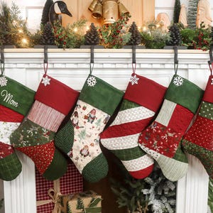 Pode incluir: Seis meias de Natal penduradas numa lareira branca. As meias têm padrões acolchoados vermelhos e verdes, com detalhes de flocos de neve e nomes personalizados. A lareira é decorada com vegetação e luzes.