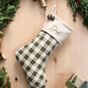 Può includere: Una calza di Natale con un motivo a quadri verdi e bianchi. La parte superiore della calza è bianco sporco con la parola "Dad" ricamata in verde. La calza è decorata con una piccola campana di metallo e decorazioni natalizie.