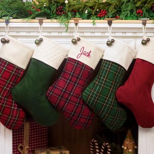 Puede incluir: Cinco calcetines de Navidad colgados en fila, con patrones alternos de cuadros rojos, verde sólido y rojo sólido. Cada calcetín tiene un puño de color crema, y el calcetín central está bordado con el nombre Jack en letra roja.