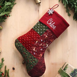 以下が含まれることがあります： 「Chloe」という名前が刺繍された赤い袖口が付いた、キルト加工のクリスマスストッキング。ストッキングは、トナカイとヒイラギのデザインが施された赤、緑、模様の生地のパッチワークセクションが特徴です。雪の結晶のチャームがループからぶら下がっています。