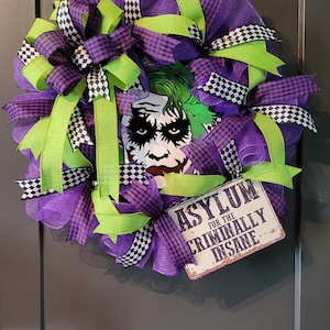 May include: A decorative wreath featuring purple mesh, lime green ribbons, and black and white checkered ribbons. In the center is a graphic of a character with green hair and face paint. A rectangular sign reads ASYLUM FOR THE CRIMINALLY INSANE.