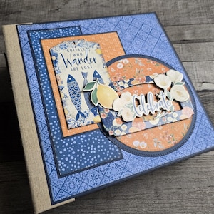 Peut inclure: Un album de scrapbooking décoratif avec un motif floral bleu et orange. L'album présente une étiquette avec le texte "Not all who wander are lost" et un embellissement "Celebrate". La couverture a un motif.