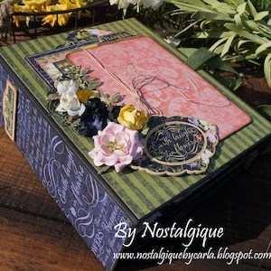 Può includere: Un album di scrapbook decorativo a tema floreale. L'album presenta una copertina a righe rosa e verdi, adornata con fiori di carta nei toni del rosa, bianco, giallo e blu. L'album è impreziosito da un'etichetta decorativa che recita "Earth laughs in flowers."