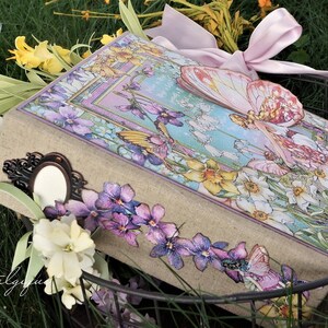 K&ouml;nnte beinhalten: Ein Scrapbook im Vintage-Stil mit einem floralen Design, das eine Fee mit Schmetterlingsfl&uuml;geln zeigt. Der Umschlag ist mit violetten Blumen, gelben Narzissen und einem rosa Band verziert.