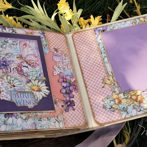 K&ouml;nnte beinhalten: Ein Scrapbook mit einem violetten und rosafarbenen Karomuster. Der Umschlag zeigt ein florales Design mit einer Fee und den Worten "Fairy Wings". Die Innenseite des Scrapbooks hat einen violetten Hintergrund mit einem floralen Design und den Worten "Make a Wish".