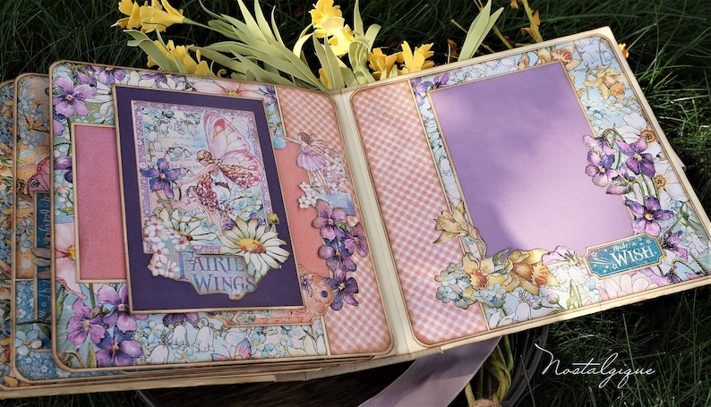 K&ouml;nnte beinhalten: Ein Scrapbook mit einem violetten und rosafarbenen Karomuster. Der Umschlag zeigt ein florales Design mit einer Fee und den Worten "Fairy Wings". Die Innenseite des Scrapbooks hat einen violetten Hintergrund mit einem floralen Design und den Worten "Make a Wish".