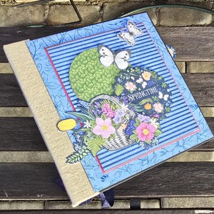 May include: A blue scrapbook with a floral design and the word "Springtime" on the front. The scrapbook has a burlap spine and a decorative butterfly on the front.