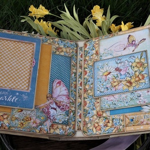 K&ouml;nnte beinhalten: Ein offenes Scrapbook mit einem floralen Design mit gelben Narzissen, Schmetterlingen und dem Text "Glaube an die Magie". Das Scrapbook hat mehrere Taschen und Rahmen f&uuml;r Fotos und Verzierungen.