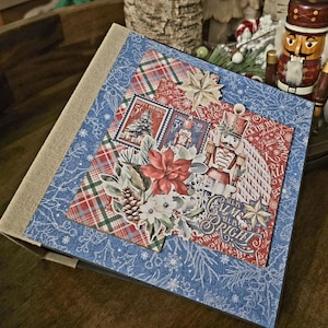 May include: A handmade scrapbook with a blue cover featuring a snowflake pattern. The scrapbook is decorated with Christmas-themed paper, including a nutcracker, poinsettias, and plaid patterns. The cover has a beige fabric spine.
