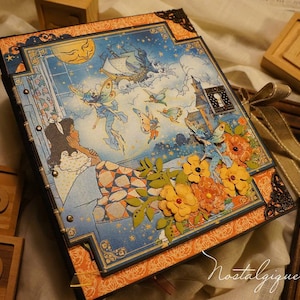 Könnte beinhalten: Ein dekoratives Scrapbook mit einem Fantasy-Themen-Cover. Das Cover zeigt eine skurrile Illustration von Feen, einem Schiff und einem Schloss vor einem Sternenhimmel. Das Scrapbook ist mit orangefarbenen floralen Verzierungen und dekorativen Eckakzenten verziert. Ein Band ist um den Buchrücken gebunden.