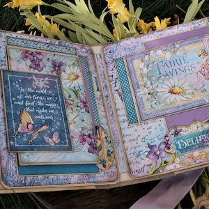 K&ouml;nnte beinhalten: Ein Scrapbook mit einem violetten, blauen und gr&uuml;nen Blumendesign. Das Scrapbook ist ge&ouml;ffnet und zeigt eine Seite mit einem Zitat &uuml;ber das Finden von Magie inmitten des Lebens. Die Seite zeigt auch eine Fee mit Fl&uuml;geln und eine G&auml;nsebl&uuml;mchen.