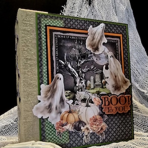 Op de afbeelding: Een Halloween-thema scrapbookalbum met een stoffen rug en een decoratieve voorkant. De voorkant toont een spookachtige scène met geesten, pompoenen en de zin "BOO! To You." De achtergrond omvat een begraafplaats en een volle maan.