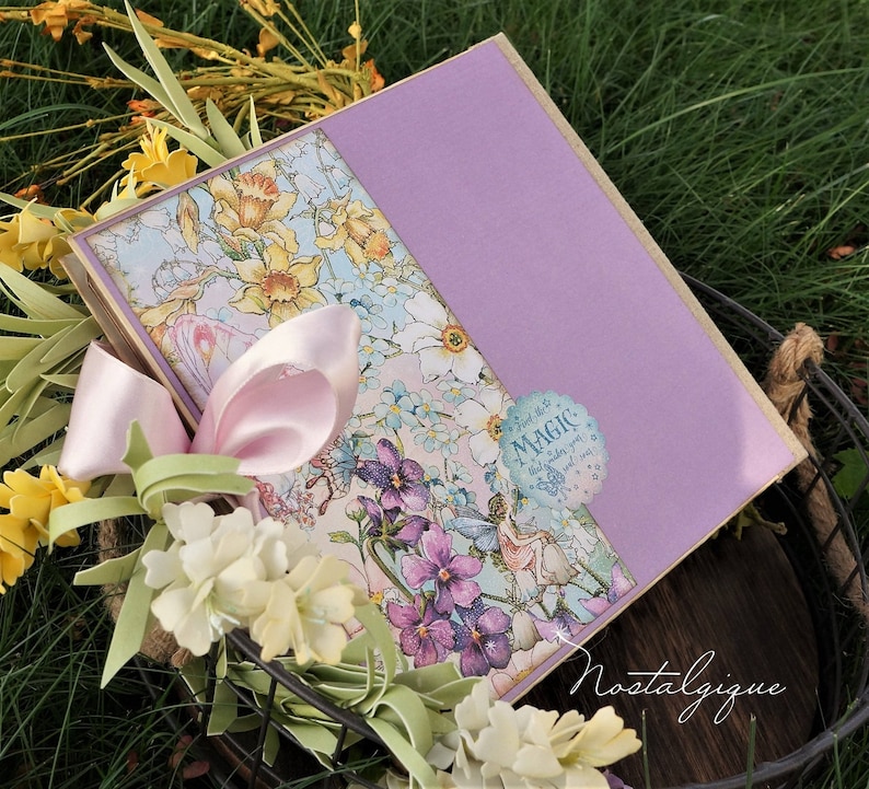 K&ouml;nnte beinhalten: Ein lilafarbener und floral verzierter Scrapbook mit einem rosa Band und einem Aufkleber mit der Aufschrift "Finde die Magie" auf dem Einband. Der Scrapbook liegt in einem Drahtkorb mit gelben Blumen.