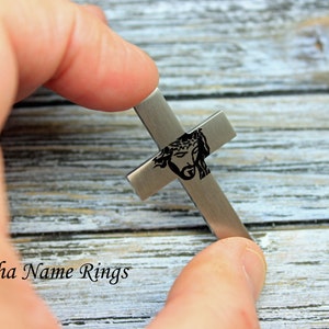 May include: A silver cross pendant with a black engraved image of Jesus Christ's face. The cross is being held by a hand.