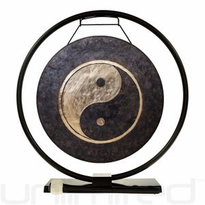 May include: A dark, round gong with a Yin Yang symbol in the center, suspended within a black metal frame. The gong is mounted on a black base. The Yin Yang symbol is a light color, contrasting with the dark gong.
