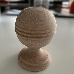 May include: A wooden finial with a round top and a base. The top has three grooves around the circumference. The finial is made of light wood.