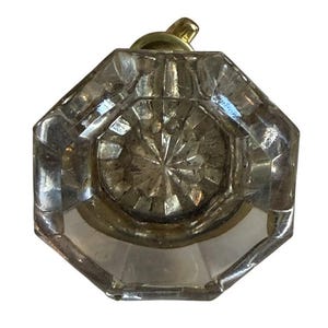 May include: Clear, faceted glass door knob with a brass-colored metal base and stem. The knob has an octagonal shape with a starburst design in the center. A small, brass-colored loop is at the top.