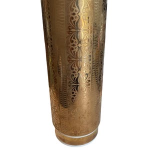 May include: Tall, cylindrical gold-colored vase with an ornate, repeating floral pattern. The vase has a white rim and base, and the gold design covers the entire surface. A decorative piece for home decor.