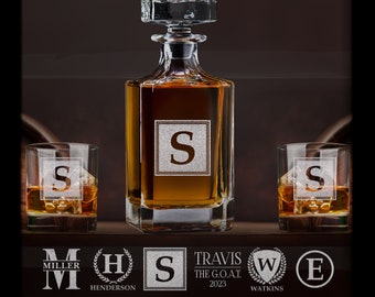 May include: A crystal whiskey decanter and two matching glasses, each engraved with the letter "S" within a square. The decanter is filled with amber liquid. The set is displayed on a wooden shelf, with various logo designs below.