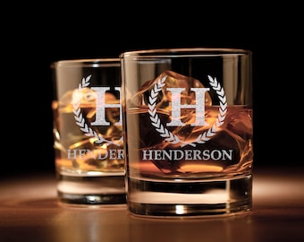 May include: Two clear glass whiskey tumblers with a silver etched design. The design features a wreath with the letter "H" in the center and the name "Henderson" below. The glasses are filled with amber liquid and ice cubes.